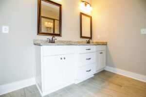 White double bathroom vanity with stone countertop, framed mirrors, dark fixtures, and wood-look tile flooring for a residential bathroom project.